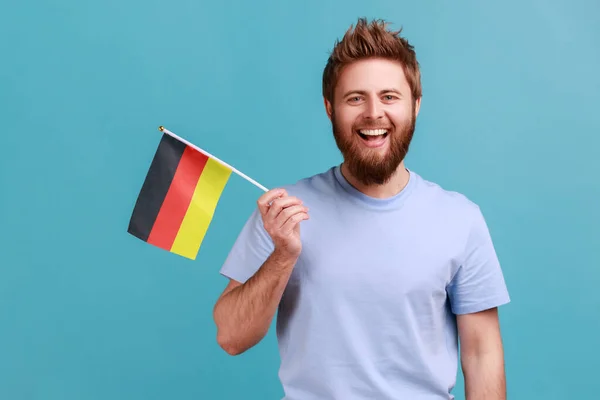 为庆祝10月3日的德国国庆日 高兴地高举德国国旗的大胡子男子的画像 表达了积极的情感 室内拍摄被蓝色背景隔离 — 图库照片