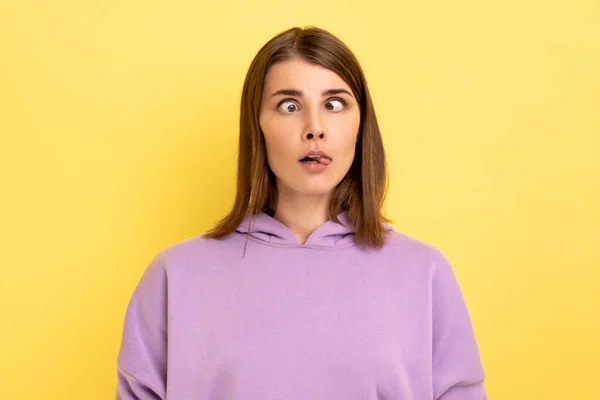 目と舌の間に立って髪を若い大人の愚かな女性の肖像画 子供っぽい行動は 紫色のパーカーを着て 黄色の背景に隔離された屋内スタジオショット — ストック写真