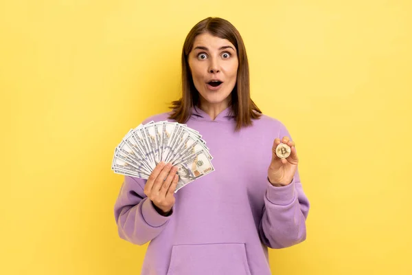 ビットコインとドル紙幣の大きなファンを保持するショックを受けた女性の肖像画 紫色のパーカーを着て 暗号通貨を交換します 黄色の背景に隔離された屋内スタジオショット — ストック写真