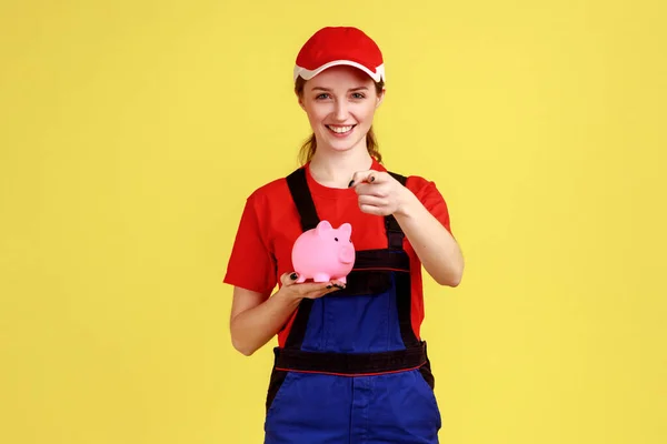 Attractive smiling handy woman standing with piggybank and pointing finger to camera, expressing positive emotions, wearing overalls and red cap. Indoor studio shot isolated on yellow background.