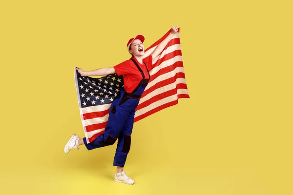 Full length portrait of excited worker woman holding with american flag in hands, pretending she is flying, wearing overalls and red cap. Indoor studio shot isolated on yellow background.