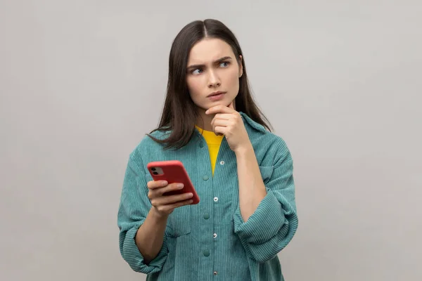 Pensive thoughtful woman thinks about answering message on smartphone and looking away, using mobile device for communication, wearing casual jacket. Indoor studio shot isolated on gray background.