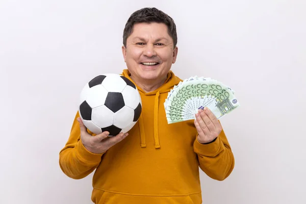 サッカーボールを示す中年の男性と100ユーロ紙幣の楽しさ笑顔 スポーツのための賭けの多くを獲得し 都市スタイルのパーカーを着て 白い背景に隔離された屋内スタジオショット — ストック写真
