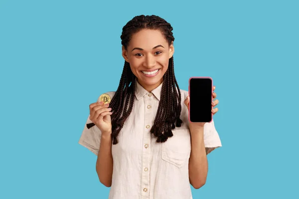 美丽的女人 带着黑发微笑 手持手机和黄金比特币 为密码应用程序做广告 穿着白衬衫 室内拍摄被蓝色背景隔离 — 图库照片