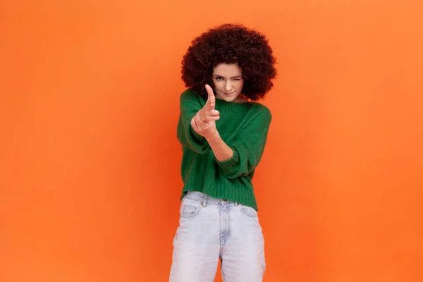 緑のカジュアルなセーターの中でアフロの髪型を持つ女性のカメラに指のピストルを指して 撮影には 不満怒っていると危険な探して脅迫 オレンジの背景に隔離された屋内スタジオショット — ストック写真