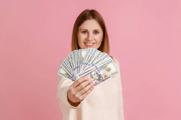 笑顔豊かな金髪の女性は 幸せな表情でカメラを見て 宝くじを獲得し 白いセーターを着て 多くのドル紙幣を保持する ピンクの背景に隔離された屋内スタジオショット — ストック写真