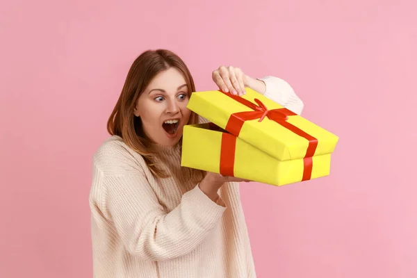 Portrait of excited young adult blond woman looking inside yellow present box, being interested what inside, wearing white sweater. Indoor studio shot isolated on pink background.