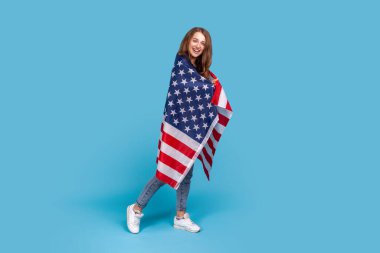 Çizgili kazak giyen, Amerikan bayrağına sarılı duran, işçi bayramını kutlayan, devletin iş desteğini kutlayan gülümseyen bir kadının tam boy portresi. Mavi arka planda kapalı stüdyo çekimi.