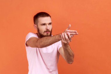 Ciddi ve cesur sakallı bir adamın parmaklarını tutup silah tutuyormuş gibi yapması, düşmanı hedef alması, nefsi müdafaa, pembe tişört giymesi. Turuncu arka planda kapalı stüdyo çekimleri.