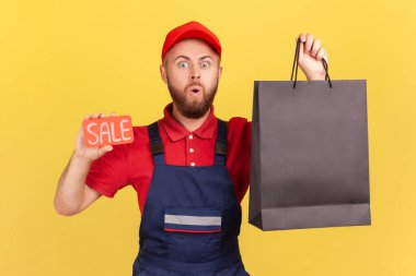 Şok olmuş kurye adamın portresi kameraya ağzı açık bakıyor, elinde siyah alışveriş çantası ve satılık sözcüğü tutuyor, mavi tulum giyiyor. Sarı arka planda kapalı stüdyo çekimleri.
