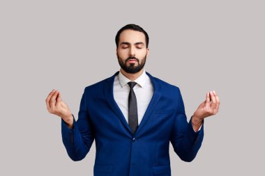 Kollarını kaldırmış, yoga yaparak meditasyon yapan, resmi stil kıyafeti giymiş rahat ve sakallı bir adamın portresi. Gri arka planda izole edilmiş kapalı stüdyo görüntüleri.