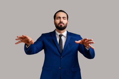 Kör sakallı bir iş adamının portresi gözleri kapalı yürüyor ve resmi bir takım elbise giyip yol bulmak için ellerini uzatıyor. Gri arka planda izole edilmiş kapalı stüdyo görüntüleri.