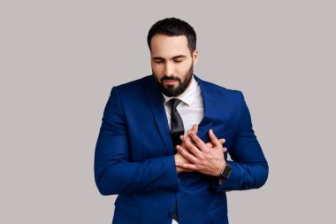 Hasta sakallı adam gözlerini kapatıyor göğsüne dokunuyor ani bir ağrı, kalp krizi riski, acil durum durumu, resmi bir takım elbise giyiyor. Gri arka planda izole edilmiş kapalı stüdyo görüntüleri.