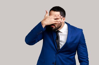 Meraklı sakallı bir adamın portresi. Gizlice parmaklarının arasından gözetliyor, sırlar arıyor, merak ediyor ve resmi bir takım elbise giyiyor. Gri arka planda izole edilmiş kapalı stüdyo görüntüleri.