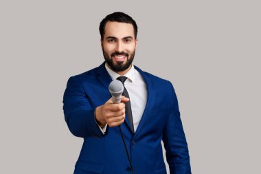 Gülümseyen sakallı bir adamın portresi mikrofon uzatıyor, sorular soruyor, röportaj yapıyor, resmi bir takım elbise giyiyor. Gri arka planda izole edilmiş kapalı stüdyo görüntüleri.