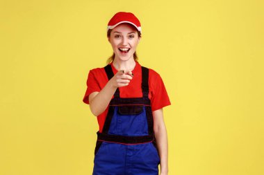 Heyecanlı mutlu işçi kadın portresi kamerayı işaret ediyor, yüksek sesle gülüyor, komik fıkralar duyuyor, tulum giyiyor ve kırmızı şapka takıyor. Sarı arka planda kapalı stüdyo çekimleri.
