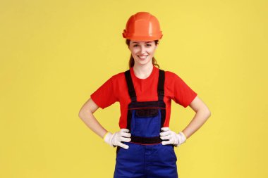 Gülümseyen, kendine güvenen, el ele tutuşan, kameraya bakan, tulum, eldiven ve koruyucu kask takan bir kadının portresi. Sarı arka planda kapalı stüdyo çekimleri.