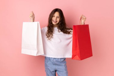 Beyaz tişört giyen mutlu küçük bir kızın portresi elinde kağıt poşetler, alışveriş merkezi indirimleri ve iyi alışverişlerle heyecanlanıyor. Pembe arka planda kapalı stüdyo çekimleri.