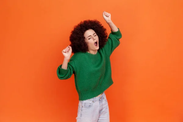Sleepy woman with Afro hairstyle wearing green casual style sweater yawning and raising hands up, feeling fatigued of overwork, open mouth. Indoor studio shot isolated on orange background.