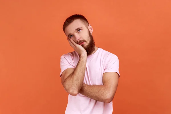 满头胡须的男人面容憔悴 面容冷漠 穿着粉色T恤 手拿着相机看着他的画像 在橙色背景下被隔离的室内拍摄 — 图库照片