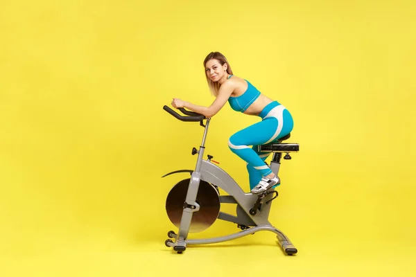 家でのスポーツ魅力的な女性のトレーニングのサイドビューの肖像画は エキサイティングな自転車を使用して カメラで笑みを浮かべて 青いスポーツウェアを着ている 黄色の背景に隔離された屋内スタジオショット — ストック写真