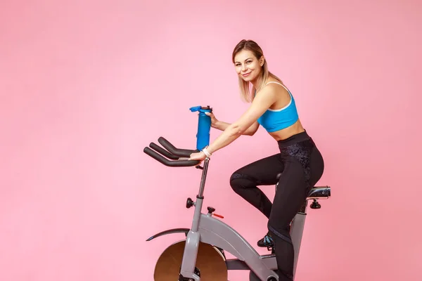 スポーツ女性のサイドビューは 運動自転車に乗って カーディオワークアウト カメラを見て 水のボトルを保持し スポーツタイツとトップを着用 ピンクの背景に隔離された屋内スタジオショット — ストック写真