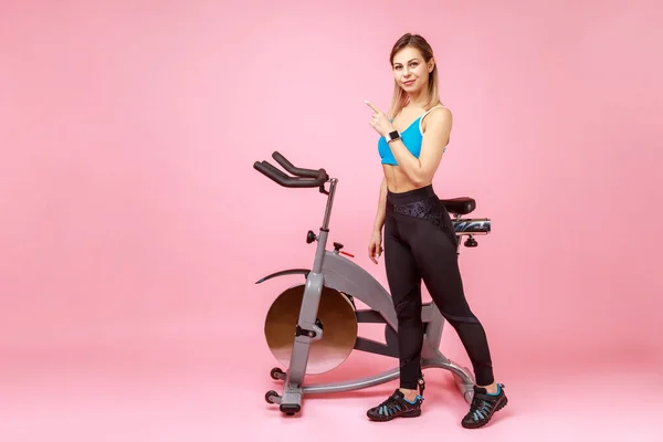 Slim athletic woman posing near exercise bike and pointing finger aside, presenting copy space for advertisement, wearing sports tights and top. Indoor studio shot isolated on pink background.