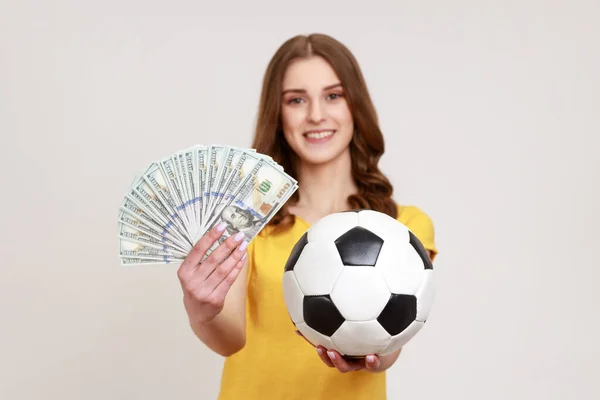 サッカーボールとドル紙幣を保持黄色のカジュアルなスタイルのTシャツを持つ幸せなティーンエイジャーの女の子の肖像画 カメラを探して 賭けと勝利 灰色の背景に隔離された屋内スタジオショット — ストック写真