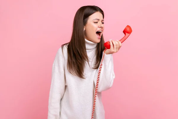 赤のレトロな携帯電話で叫んで怒っている若いブルネットの女性は 攻撃性 迷惑なクライアントを表現し 白いカジュアルなスタイルのセーターを着ています ピンクの背景に隔離された屋内スタジオショット — ストック写真