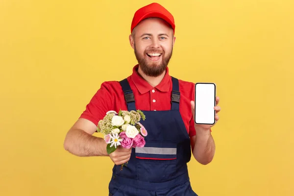 Smiling positive man courier standing with flowers in hands and showing cell phone with empty screen for advertisement, looking at camera. Indoor studio shot isolated on yellow background.
