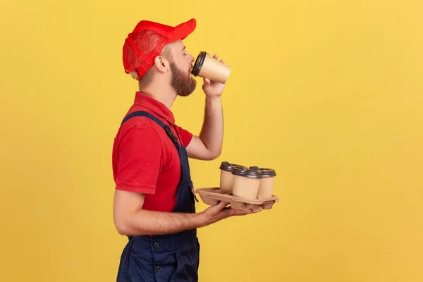 Side view portrait of bearded handy man standing with disposable cups and drinking hot beverage, needs energy for continue working. Indoor studio shot isolated on yellow background.