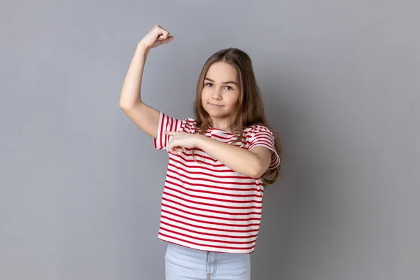 ストライプのTシャツを着た可愛い女の子の肖像画は 目標を達成するために手を上げ バイクを指し 力とエネルギーを感じています 灰色の背景に隔離された屋内スタジオショット — ストック写真