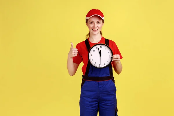 Portrait of worker woman standing with big wall clock in hands, looking at camera with smile and showing thumb up, wearing overalls and red cap. Indoor studio shot isolated on yellow background.