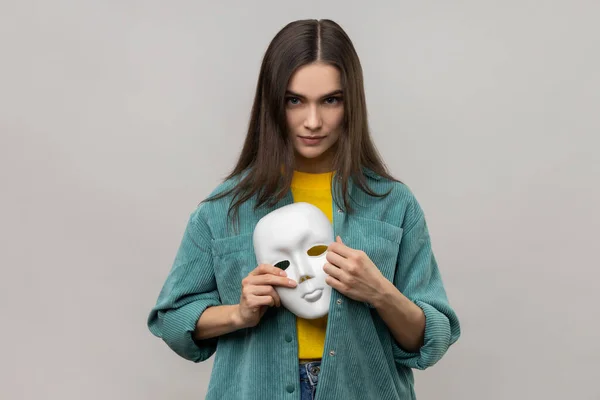 Portrait of serious bossy woman holding white mask in her hand, changing personality, hiding mask in the bosom, wearing casual style jacket. Indoor studio shot isolated on gray background.