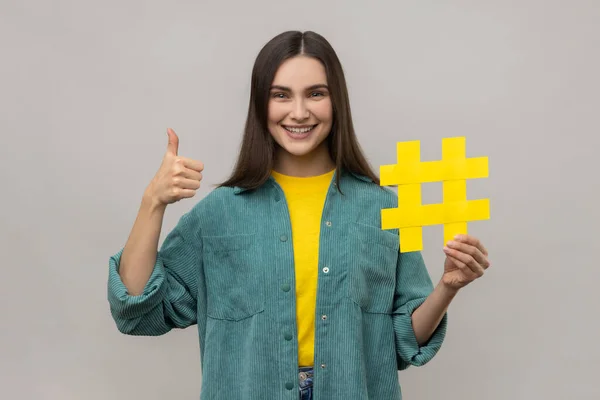 トレンドをフォローし 人気のブログ ソーシャルメディアのハッシュタグのシンボルと親指をジェスチャーを保持する楽観的な女性は カジュアルなスタイルのジャケットを身に着けている 灰色の背景に隔離された屋内スタジオショット — ストック写真