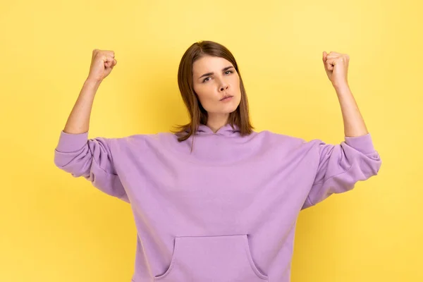 Portrait of confident strong woman, standing with strong gesture and looking at camera with serious expression, raised arms, wearing purple hoodie. Indoor studio shot isolated on yellow background.