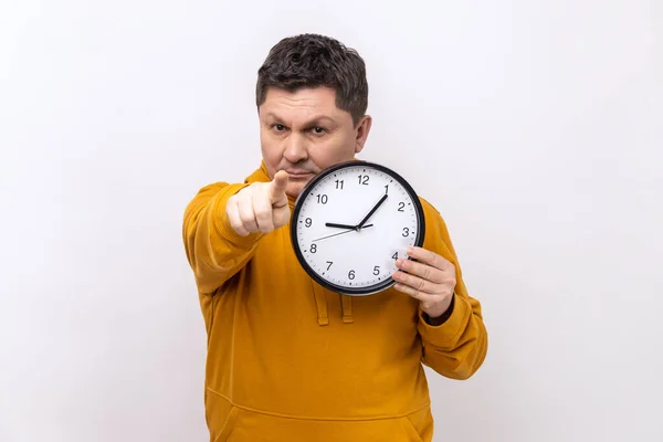 Portrait of happy positive man holding big wall clock and pointing to camera with pleasant smile, time to go, wearing urban style hoodie. Indoor studio shot isolated on white background.
