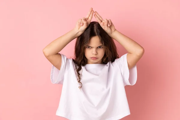 Portrait of aggressive bully little girl wearing white T-shirt showing bull horns gesture over head, frowning as before attack. Indoor studio shot isolated on pink background.