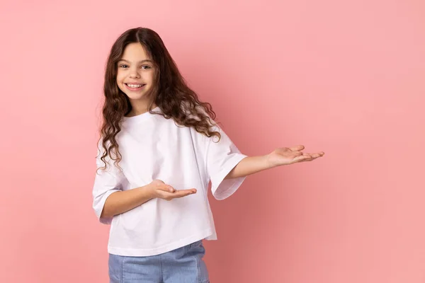 あなたの最高の広告のための壁に歓迎のジェスチャーと空のスペースを示す白いTシャツを着て優しい寛大な少女の肖像画 ピンクの背景に隔離された屋内スタジオショット — ストック写真