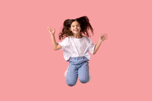 穿着白色T恤衫的积极的小女孩高兴地在空中高高地跳着 充满了活力和活力 在粉色背景下被隔离的室内拍摄 — 图库照片