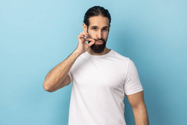 Kimseye anlatmam. Gözü korkan, beyaz tişörtlü, fermuarlı dudaklı, kameraya bakan, ağzı kapatan ve sır tutacağına söz veren bir adamın portresi. Mavi arka planda kapalı stüdyo çekimi.