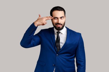Sakallı adamın portresi, parmaklarını kafasının yanında tutuyor, silahını taklit ediyor, kameraya paniklemiş bir ifadeyle bakıyor, resmi stil elbisesi giyiyor. Gri arka planda izole edilmiş kapalı stüdyo görüntüleri.