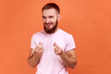 Yakışıklı genç sakallı bir adamın resmi ayakta durup kameraya parayla bakıyor ya da pembe tişört giyen İtalyan jestleri yapıyor. Turuncu arka planda kapalı stüdyo çekimleri.