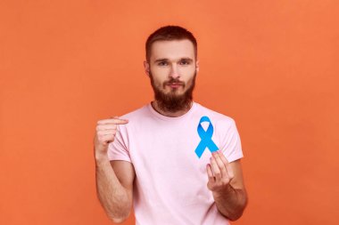 Ciddi sakallı bir adamın portresi mavi farkındalığı işaret ediyor, hastalık sembolü, kameraya bakıyor, destek oluyor, pembe tişört giyiyor. Turuncu arka planda kapalı stüdyo çekimleri.