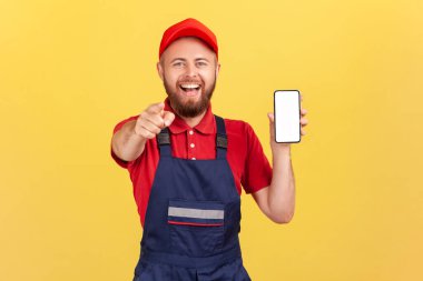 Gülümseyen mutlu işçi, mavi tulumlu, kırmızı tişörtlü, seni işaret eden ve akıllı telefonu boş ekran, online servis sipariş uygulaması ile gösteren. Sarı arka planda kapalı stüdyo çekimleri