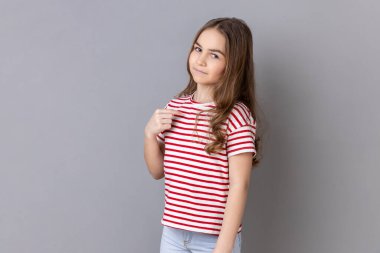 This is me. Portrait of little girl wearing striped T-shirt pointing herself and looking selfish egoistic haughty, feeling proud of own achievement. Indoor studio shot isolated on gray background. clipart