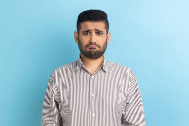 Sakallı üzgün işadamı, üzgün bir suratla kameraya bakıyor. Üzüntüsünü dile getiriyor. Kötü bir ruh hali içinde, çizgili gömlek giyiyor. Mavi arka planda kapalı stüdyo çekimi.