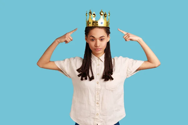Serious independent woman with black dreadlocks points at gold crown, looking with arrogance and confidence, privileged status, wearing white shirt. Indoor studio shot isolated on blue background.