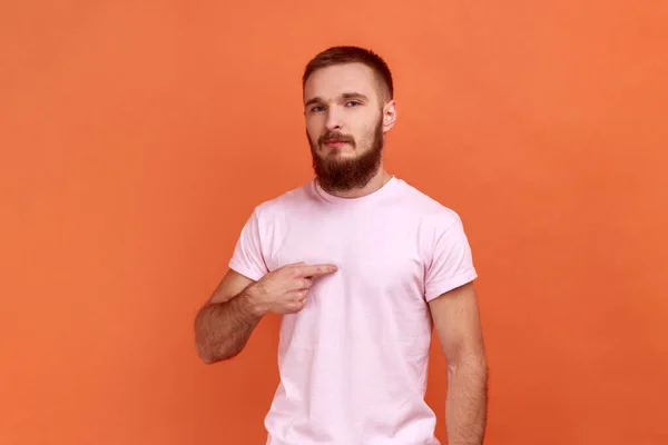 Portrait of bearded man pointing himself and looking with arrogant selfish expression, feeling successful and self-important, wearing pink T-shirt. Indoor studio shot isolated on orange background.