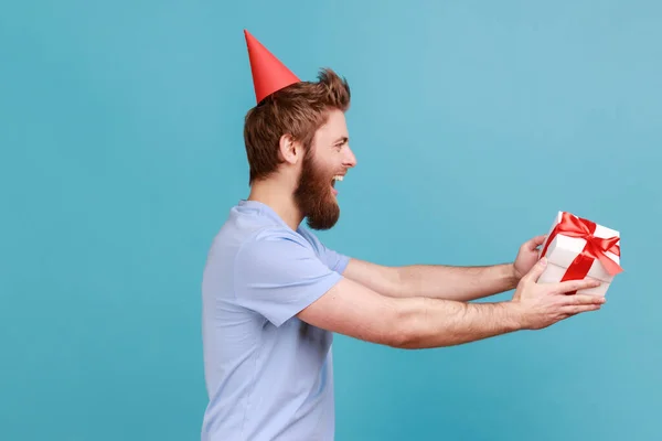 Side view of man in party cone giving birthday gift and smiling, sharing present, donating and offering surprise in box, congratulation on anniversary. Indoor studio shot isolated on blue background.